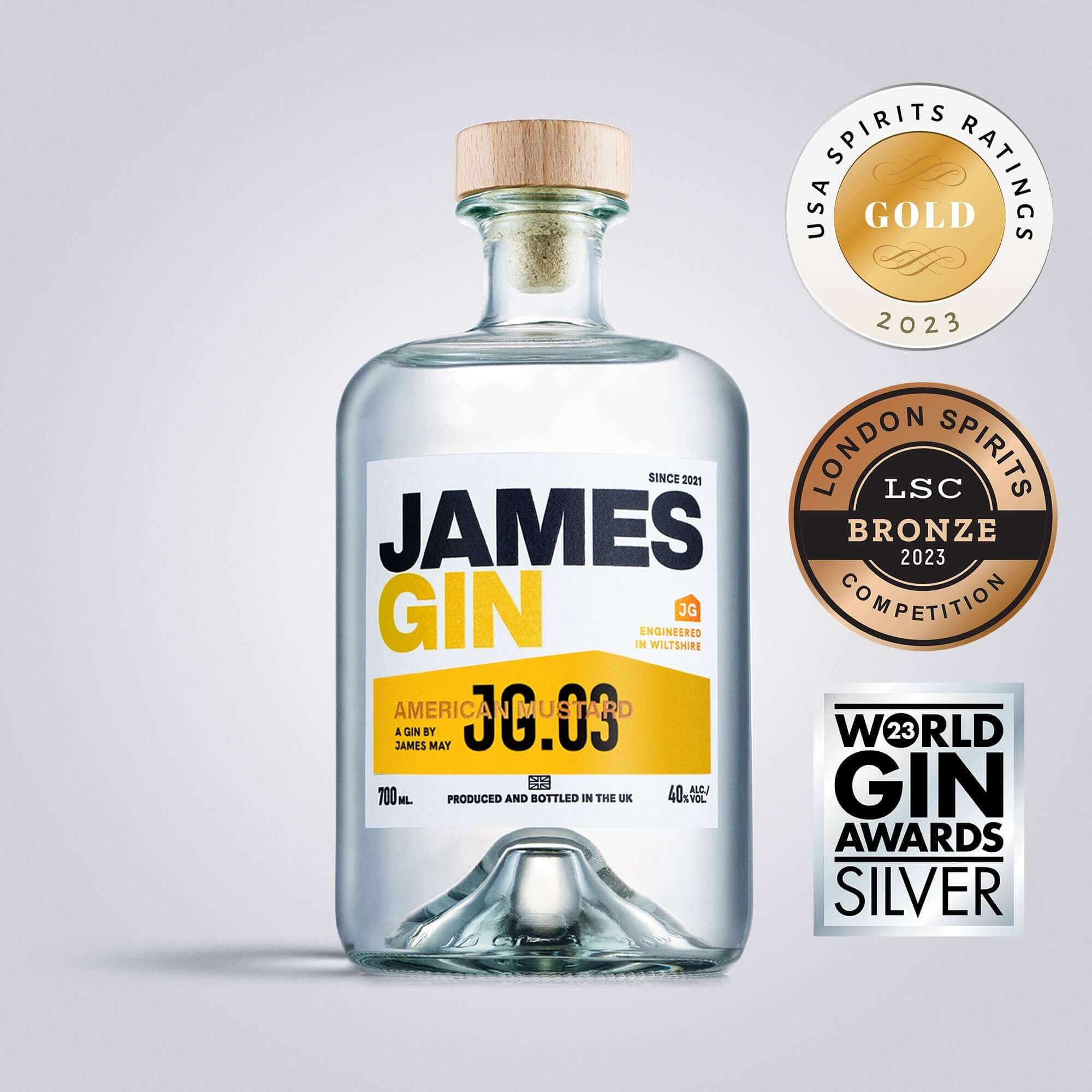 A clear 700mL bottle of James Gin American Ramstud with a wooden cap and striking yellow-blue label is shown, alongside three award badges (Gold, Silver, Bronze) and a souvenir gift key chain.