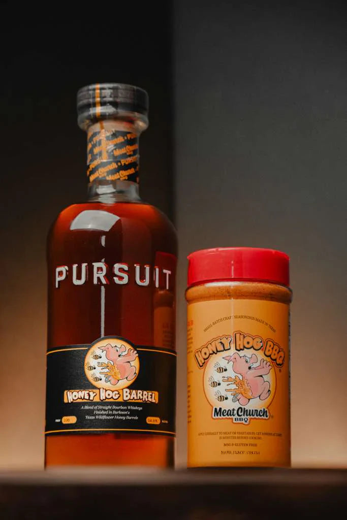 A bottle of Pursuit Spirits Pursuit x Meat Church Honey Hog Barrel Bourbon stands next to Meat Church Honey Hog BBQ seasoning, both displayed on a dark background with warm lighting.