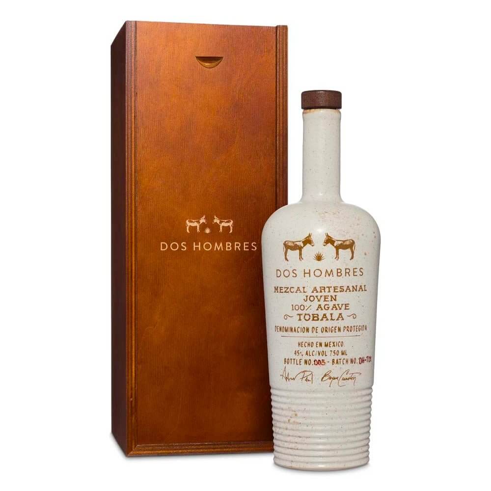 A white bottle of Dos Hombres Tobala Mezcal Limited Edition by Aaron Paul & Bryan Cranston, adorned with gold text and two donkeys, stands next to its wooden box—showcasing this exclusive release from the Dos Hombres brand.