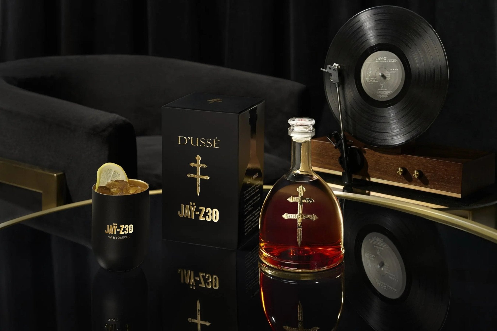 A bottle of D’USSE VSOP Jay-Z30 Limited Edition Cognac (PRE-ORDER) stands in a branded black box beside a glass with a lemon garnish, on a reflective table. A record player and velvet chair in the background add to the French cognac’s luxurious ambiance.