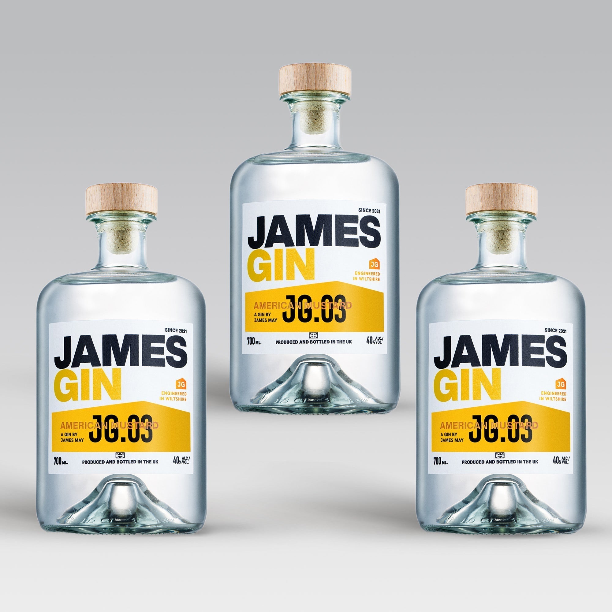 Three bottles of James Gin US "The Big, Beautiful Bundle" with wooden caps are arranged in a triangle on a light gray background. Each features a white label with bold black and yellow text, plus a souvenir gift key chain.