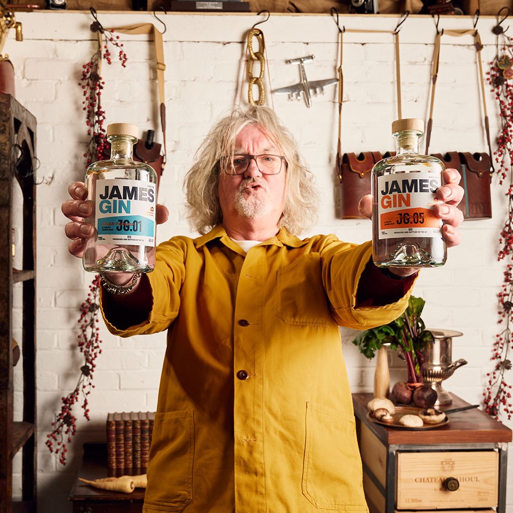 A gray-haired man in glasses and a yellow jacket stands indoors, holding two bottles of James Gin The Internationalist W/ Souvenir Gift Key Chain by James Gin US. Tools, hanging herbs, and vintage decor are visible in the background.