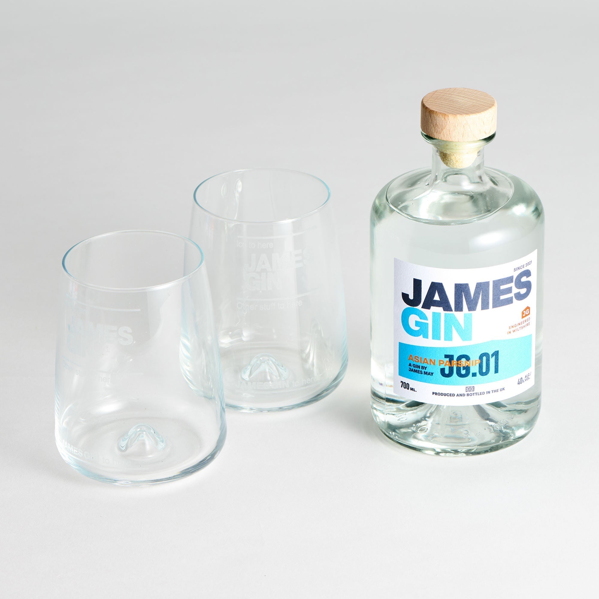 A James Gin The Sharing Cocktail Kit W/ Souvenir Gift Key Chain by James Gin US sits beside two empty clear glasses on a white surface. The bottle has a wooden cap and features a white and blue label with text and numbers.