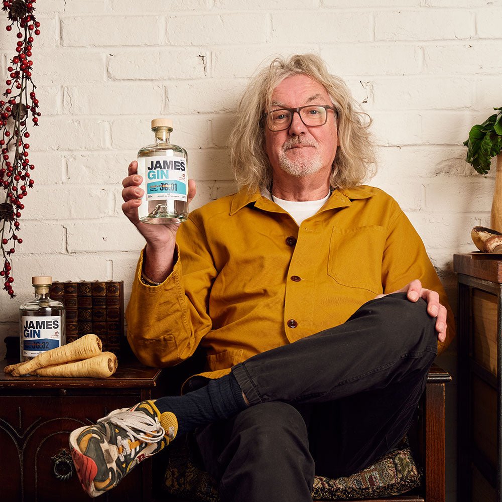 A man with gray hair and glasses in a mustard-yellow jacket and colorful sneakers sits cross-legged indoors, smiling at the camera while holding James Gin US The Six Pack Asian Parsnip & Navy Strength with a souvenir gift key chain. Another bottle is nearby.
