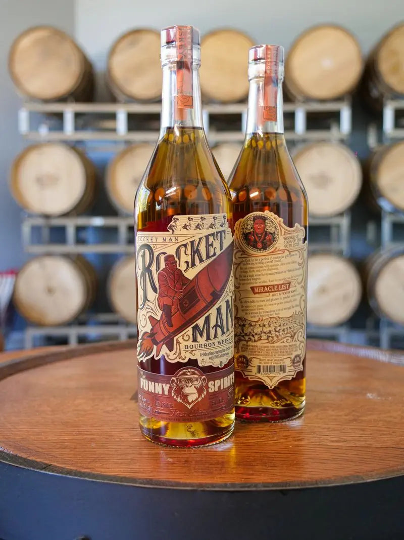 Two bottles of Rocket Man Bourbon Whiskey by Rocket Man, featuring limited release illustrated labels, rest on a barrel with rows of barrels in the background—an ideal scene for this political parody whiskey.