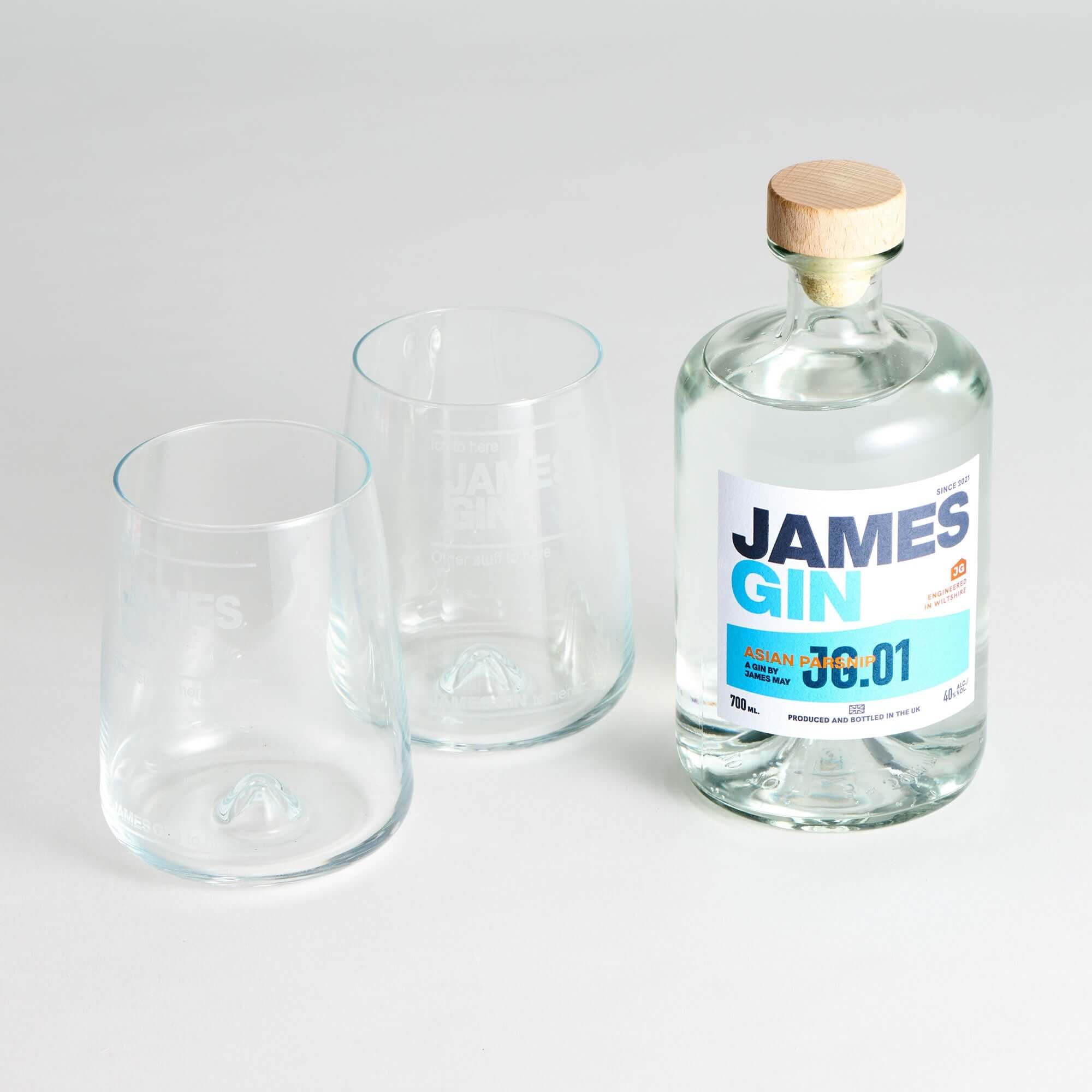 A James Gin The Sharing Cocktail Kit W/ Souvenir Gift Key Chain by James Gin US sits beside two empty clear glasses on a white surface. The bottle has a wooden cap and features a white and blue label with text and numbers.
