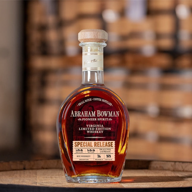 A bottle of A. Smith Bowman Distillery Abraham Bowman Special Release #26 Virginia Rye Whiskey with a wooden cork sits on wood, blurred barrels behind it. The label notes its limited edition status.