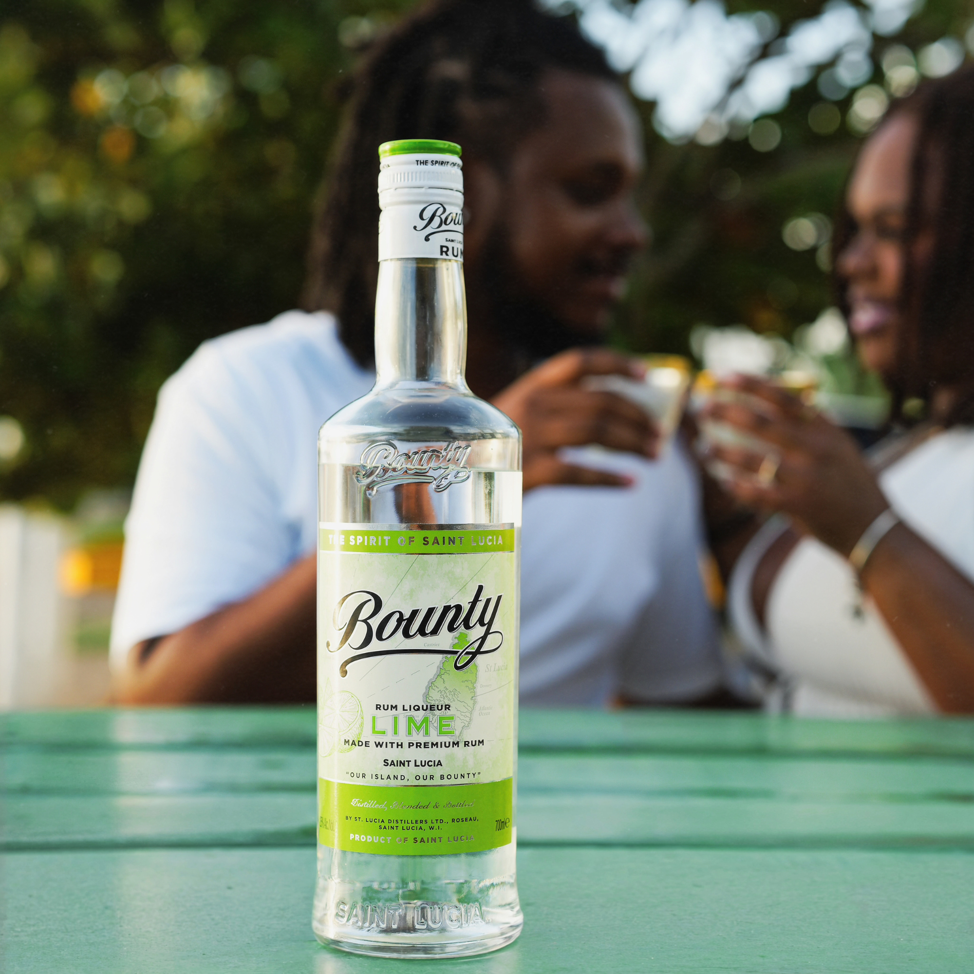 A bottle of Bounty Rum Lime 750 ml is in sharp focus on a green table, its zesty flavor from Bounty Rum perfect for long, refreshing drinks. In the blurred background, a smiling couple in white enjoys drinks and chats outdoors with trees behind them.