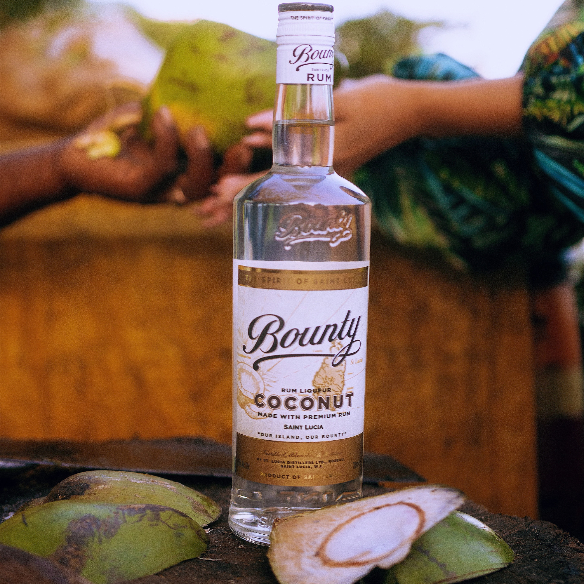 A bottle of Bounty Rum Coconut 750 ml stands on a wooden surface surrounded by opened coconuts, while blurred hands holding coconuts appear in the background.
