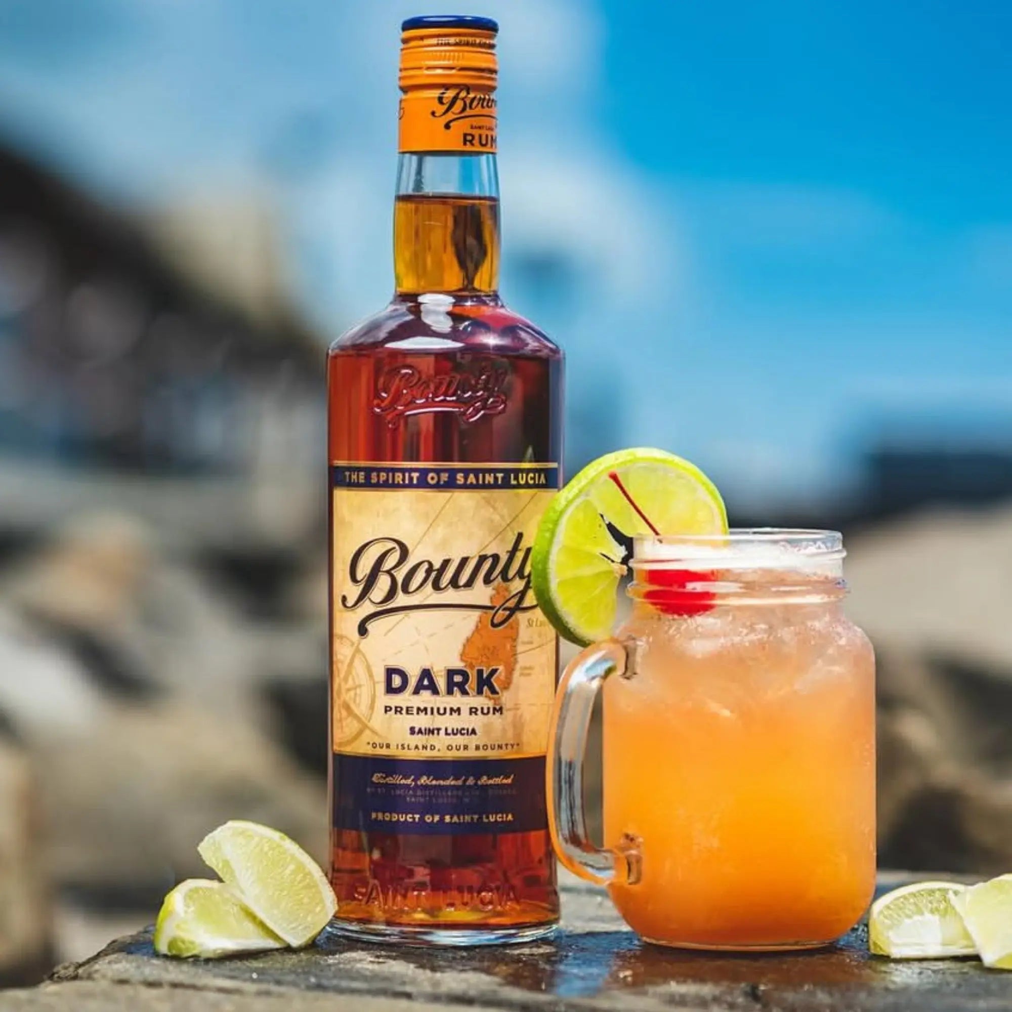 A bottle of Bounty Rum Premium Dark 750 ml stands beside a mason jar glass holding an orange cocktail garnished with a lime wheel and cherry, plus lime wedges on a rocky outdoor surface—ideal for enjoying its silky smooth mouthfeel.
