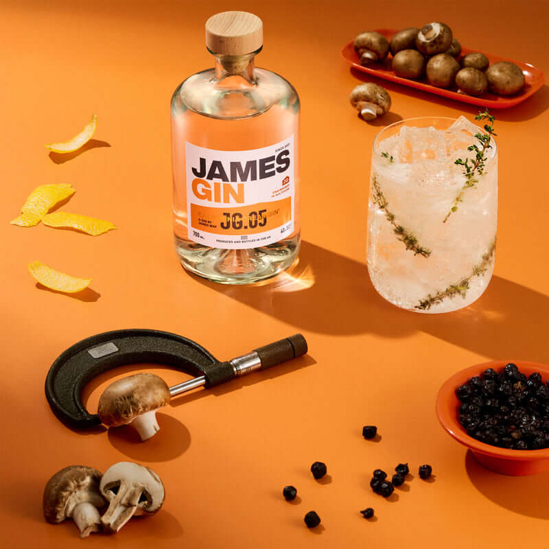 A bottle of James Gin California Dreamgin' W/ Souvenir Gift Key Chain, a glass with ice and herbs, mushrooms, orange peels, black peppercorns, a mushroom clamp, and small bowls rest on an orange surface. Enjoy botanical flavors by James Gin.