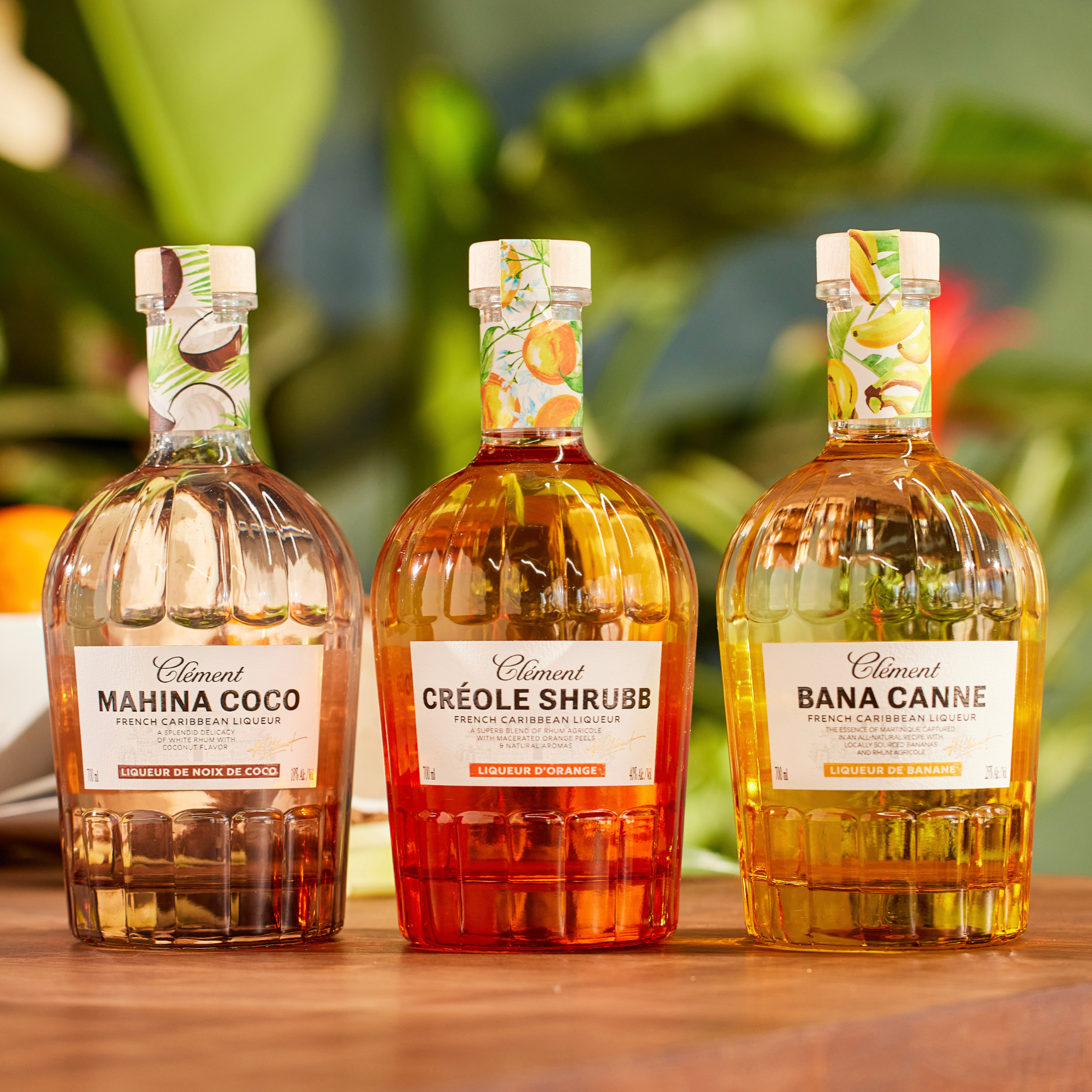 Three bottles from Rhum Clément—including the Mahina Coco Coconut Liqueur—stand side by side on a wooden surface, with Mahina Coco, a rum-based coconut liqueur, set against a blurred backdrop of lush green plants.