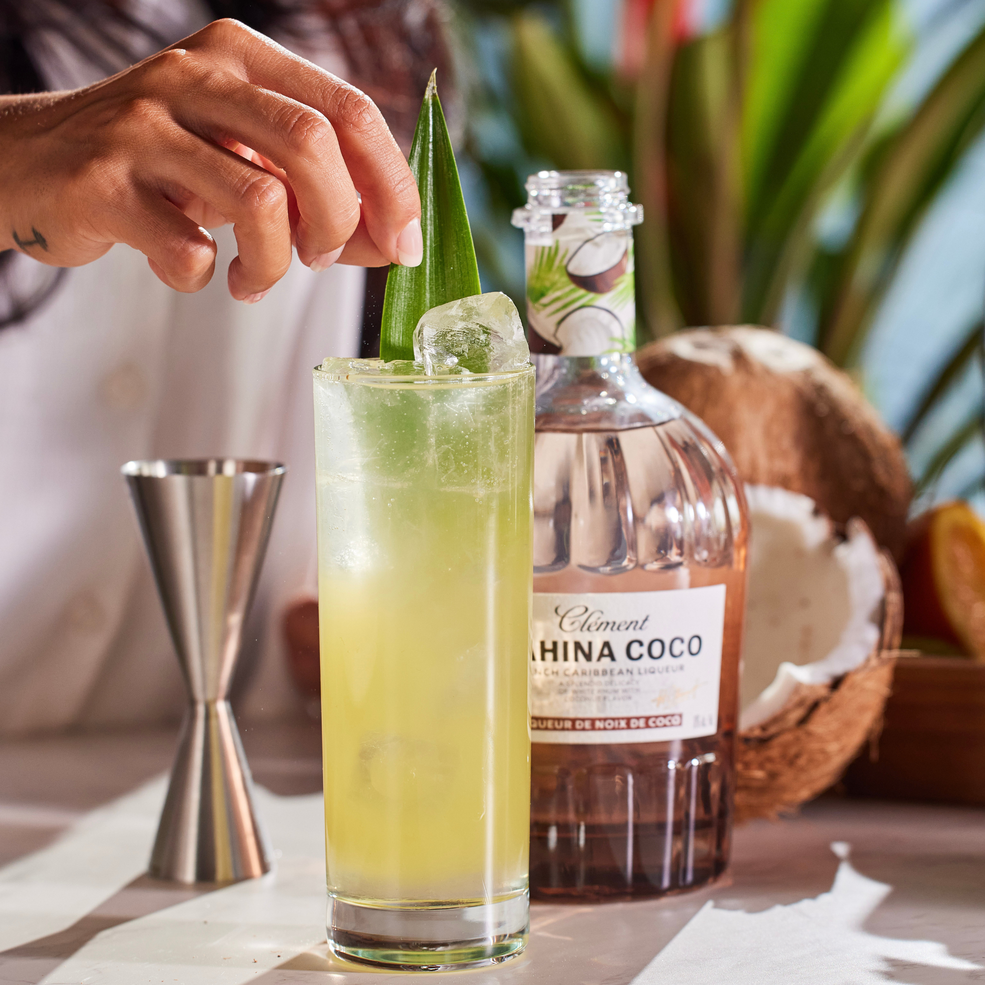 A hand adds a leaf garnish to a tall glass of yellow-green cocktail with ice. Behind it sits a bottle of Rhum Clément Mahina Coco | Coconut Liqueur from Rhum Clément, a jigger, and half a coconut on a sunlit table.