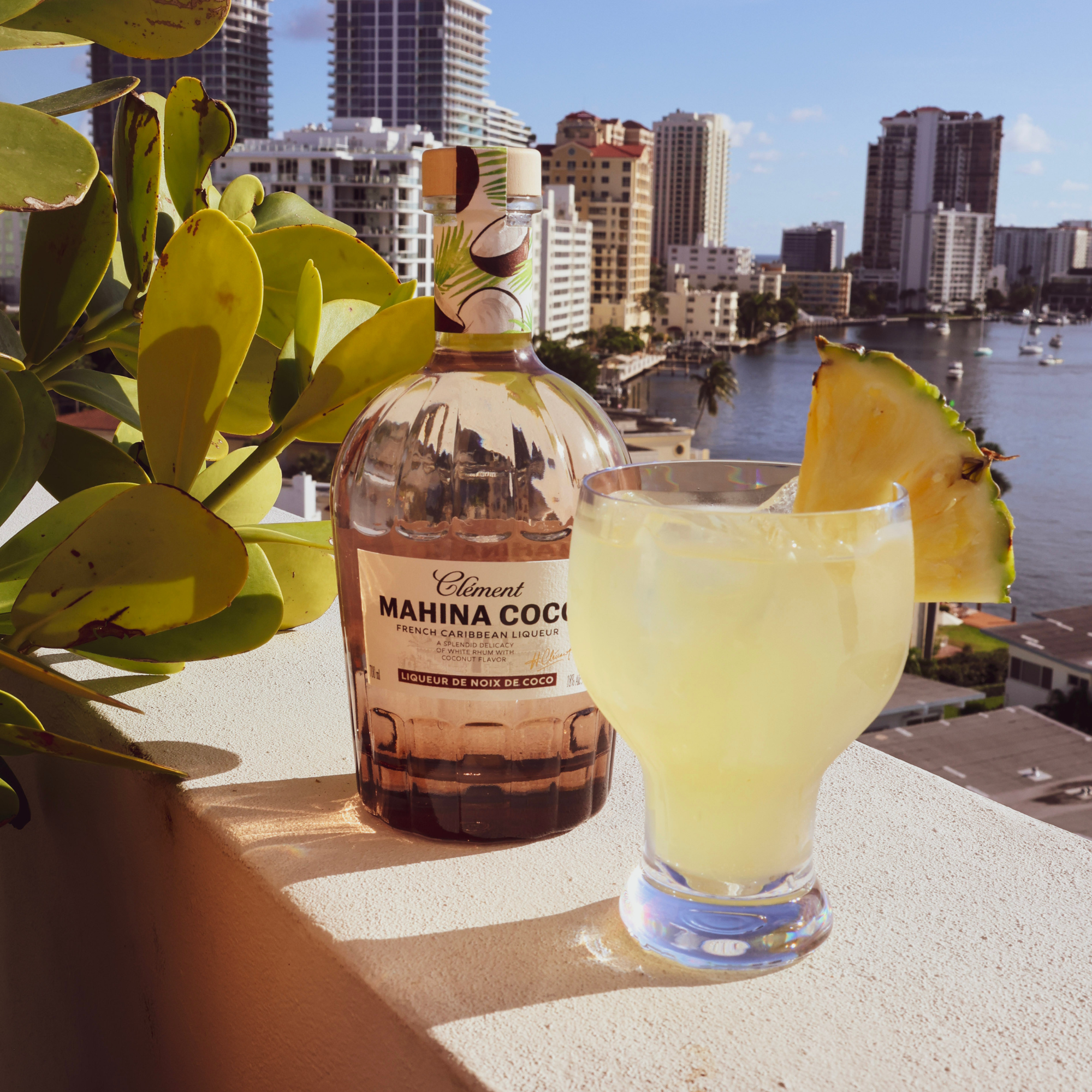 A bottle of Rhum Clément Mahina Coco Coconut Liqueur and a tropical cocktail with pineapple garnish rest on a balcony ledge, overlooking city buildings, water, and blue sky in the background.