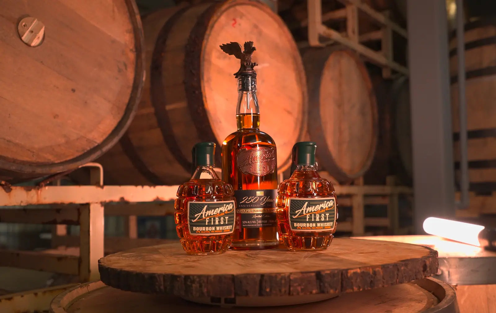 Three bottles from the America First Limited Edition Bourbon Bundle—featuring the Anniversary Bottle and two Grenade Whiskeys—are displayed on a wooden barrel, with large aging barrels in the background, creating a warm distillery setting reminiscent of classic America First Grenade Whiskey Bottles.