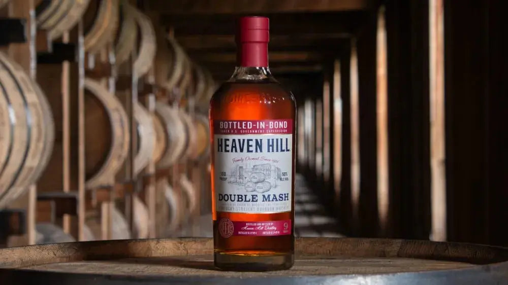 A bottle of Heaven Hill Double Mash Bottled in Bond Bourbon from Heaven Hill Distillery rests atop a barrel inside a dimly lit rickhouse, with rows of wooden barrels softly blurred in the background.