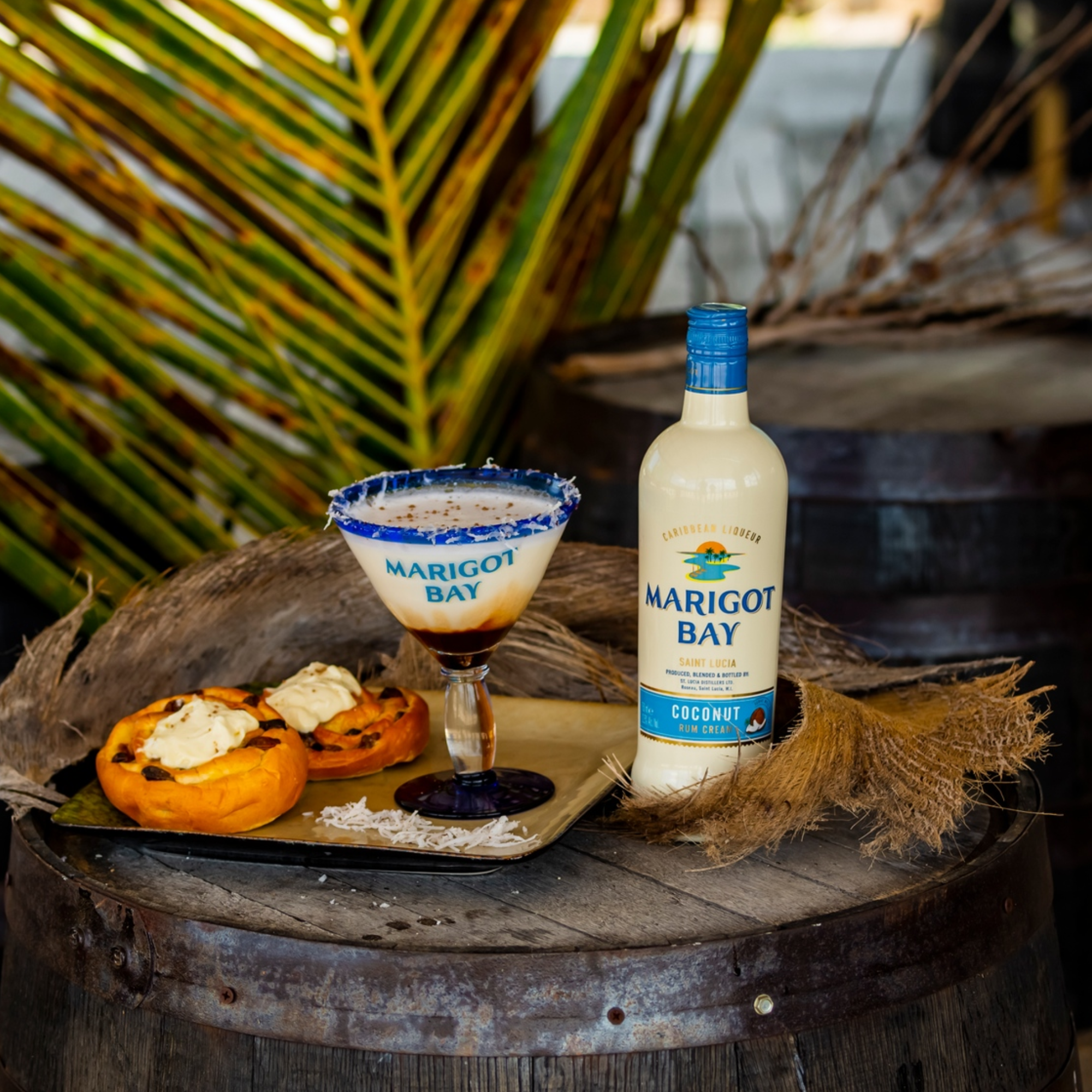 A bottle of Marigot Bay Rum's Coconut Rum Cream Liqueur, a blue sugar-rimmed cocktail, and two cream cheese pastries sit on a rustic barrel, capturing the tropical essence of Saint Lucian rum among palm leaves.