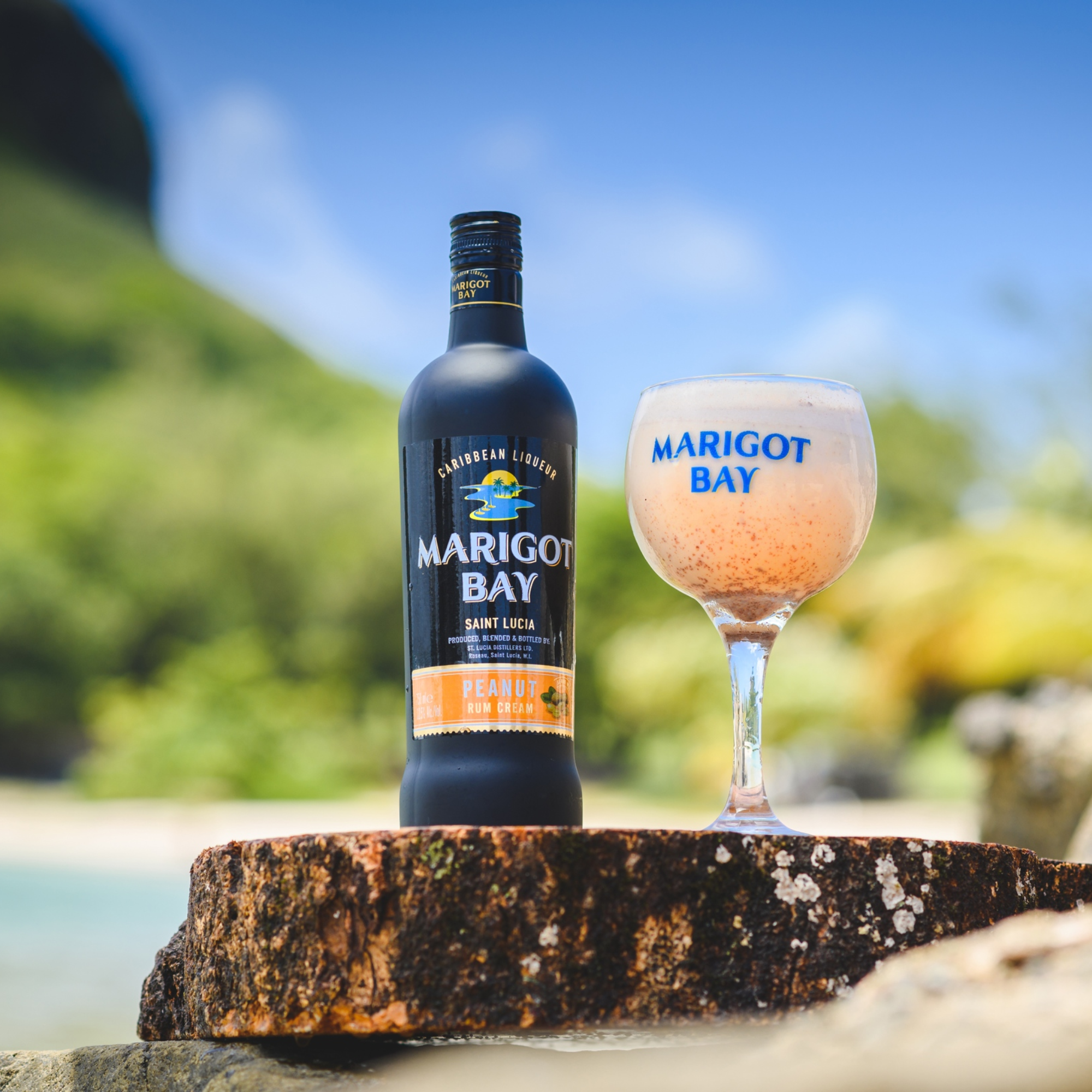 A bottle of Marigot Bay Peanut Rum Cream Liqueur by Marigot Bay Rum and a glass of the liqueur sit on a tree stump outdoors, with a tropical beach and lush greenery softly blurred in the background.