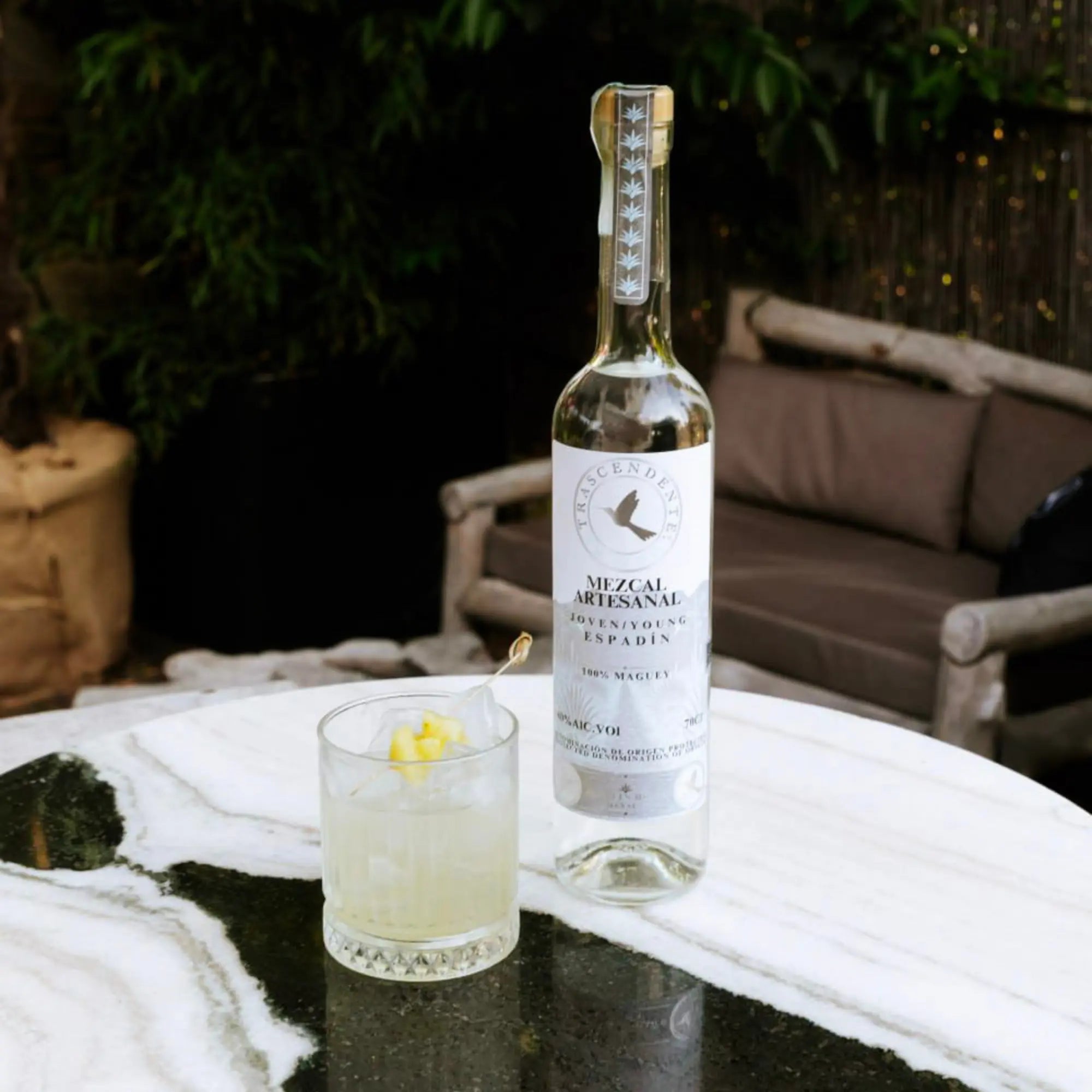A bottle of Mezcal Trascendente Espadin 700ml and a cocktail glass with citrus peel rest on a round white marble table outdoors, exuding a fresh citric aroma, with plants and a grey outdoor sofa in the background.