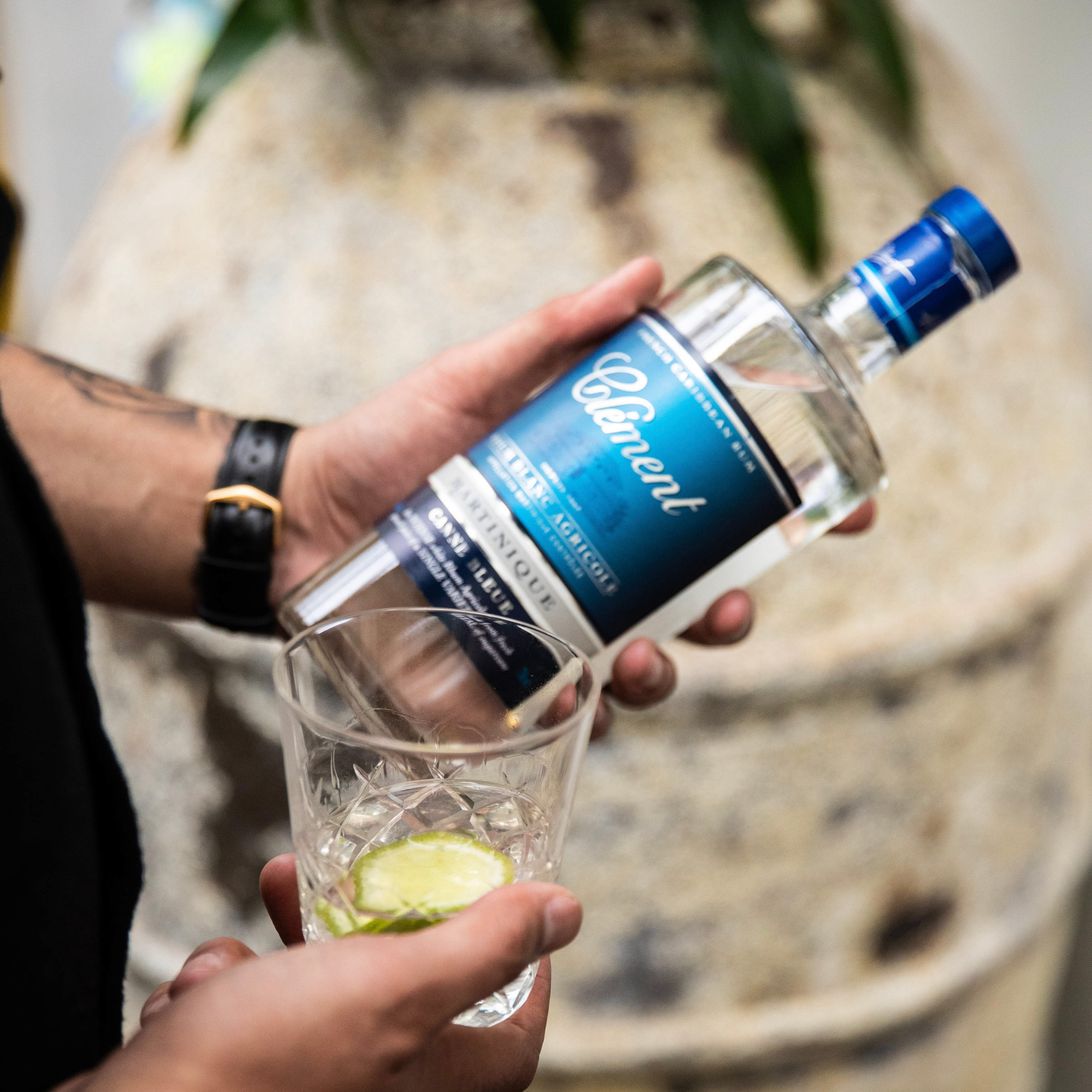 A person pours Rhum Clément Canne Bleue White Rum into a glass with lime slices. Wearing a black watch, they stand before a stone surface and greenery—an inviting scene for savoring Rhum Clément from Martinique.