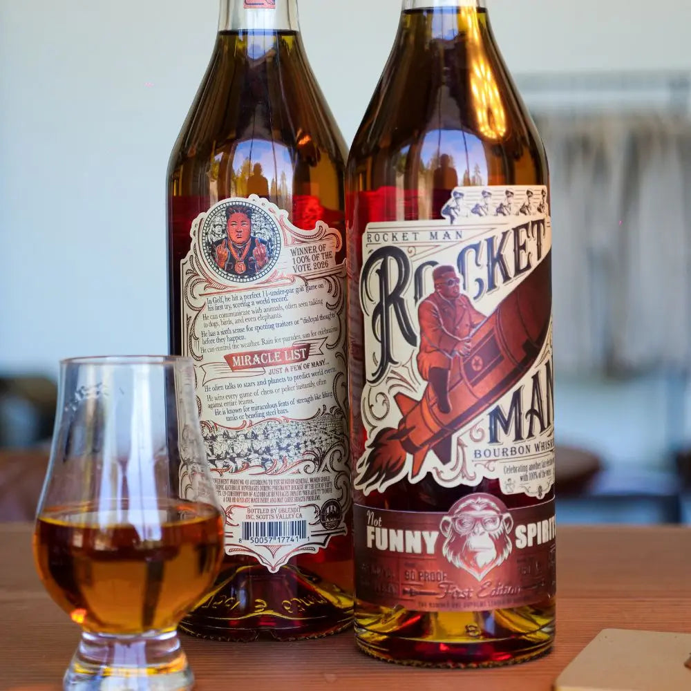 Two bottles of Rocket Man Bourbon Whiskey by Rocket Man are displayed—one featuring the front label with a man riding a rocket, the other the back. A glass of this limited release bourbon whiskey sits on a wooden surface in front.