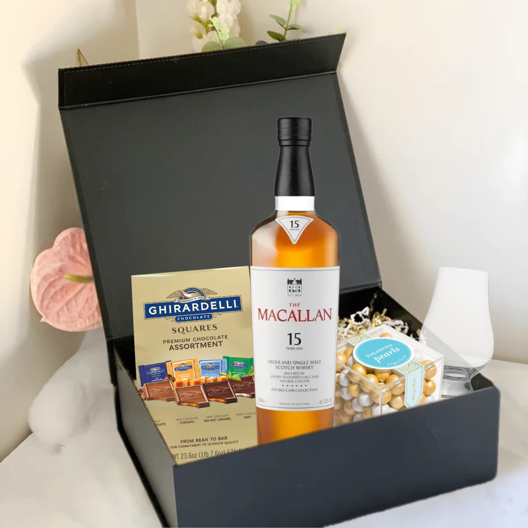 The Premium Scotch Gift Box by Main Street Liquor features The Macallan 15 Scotch, Ghirardelli chocolates, a glass, mixed nuts, and gold-wrapped treats—stylishly presented on white with pink flowers.