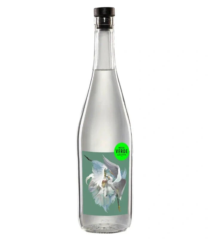 A clear glass bottle with a black cap, filled with clear liquid. The label shows a white flower on green and a small green circle reading MEZCAL VERDE. Product: Amaras Mezcal Verde Artist Series 2024 by Amaras.