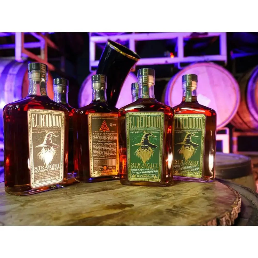 Five bottles of Eldenwood Straight Bourbon Whiskey with Drinking Horn are arranged on a wooden barrel, with large aging barrels in the background, in a dimly lit distillery setting—a scene that perfectly captures the allure of an exclusive Eldenwood bourbon gift set.