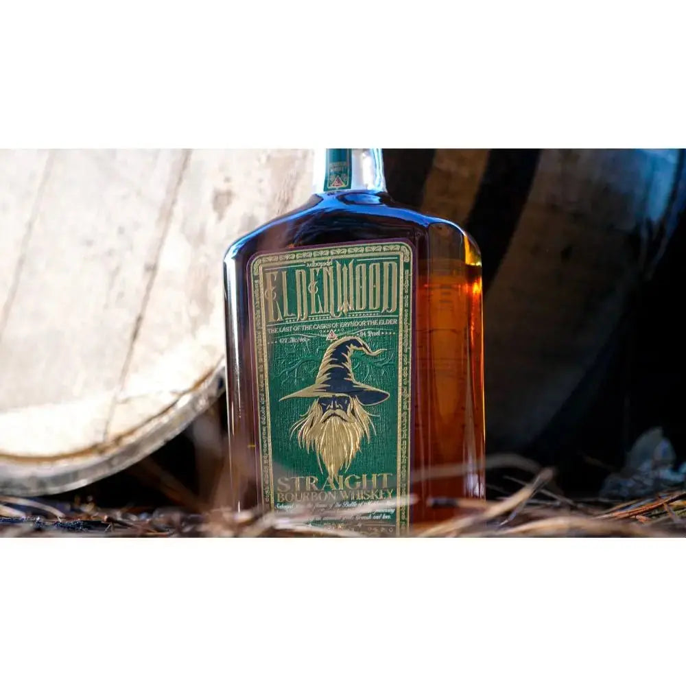 A bottle of Eldenwood Straight Bourbon Whiskey with Drinking Horn from Eldenwood, labeled Lost Woods and featuring an illustration of a wizard, is set in front of a wooden barrel and surrounded by straw—making it a perfect addition to any bourbon gift set.