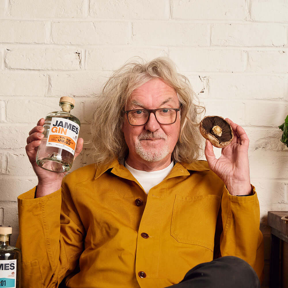 A man with gray, curly hair and glasses, in a mustard jacket, sits before a white brick wall holding a James Gin US The Internationalist bottle with souvenir gift key chain in one hand and a large portobello mushroom in the other.