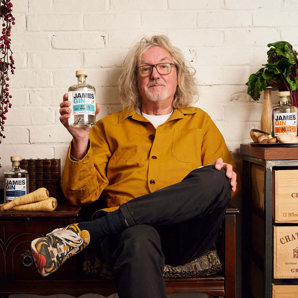 A man with shoulder-length gray hair, glasses, and a yellow jacket sits cross-legged, holding a James Gin Asian Parsnip 700mL 80 Proof with Souvenir Gift Key Chain. Other James Gin bottles and wooden crates rest nearby against a white brick wall.