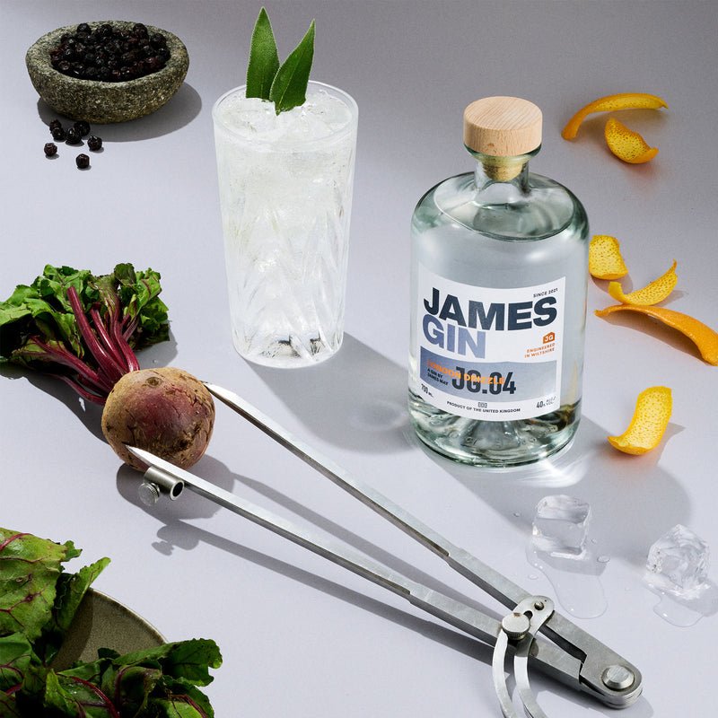 A bottle of James Gin London Drizzle 700mL 80 Proof with Souvenir Gift Key Chain, a crystal cocktail glass with mint, artisan botanicals, beetroot, orange peels, ice cubes, and spices all arranged on a white surface.