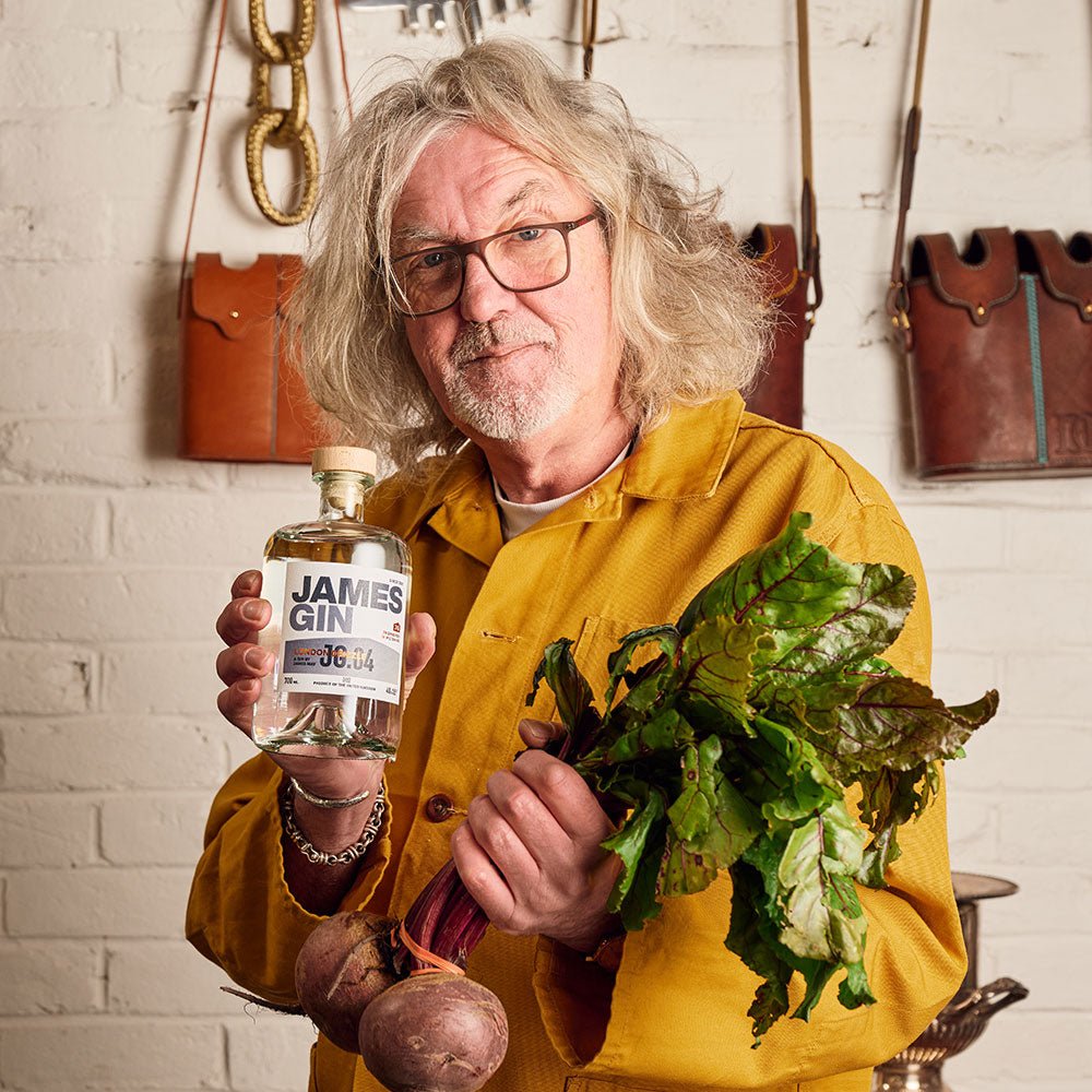 A man with long gray hair and glasses, in a yellow jacket, stands by a white brick wall with leather bags. He holds fresh beetroots in one hand and a James Gin US The Survivalist 6 Pack w/ Souvenir Gift Key Chain in the other.