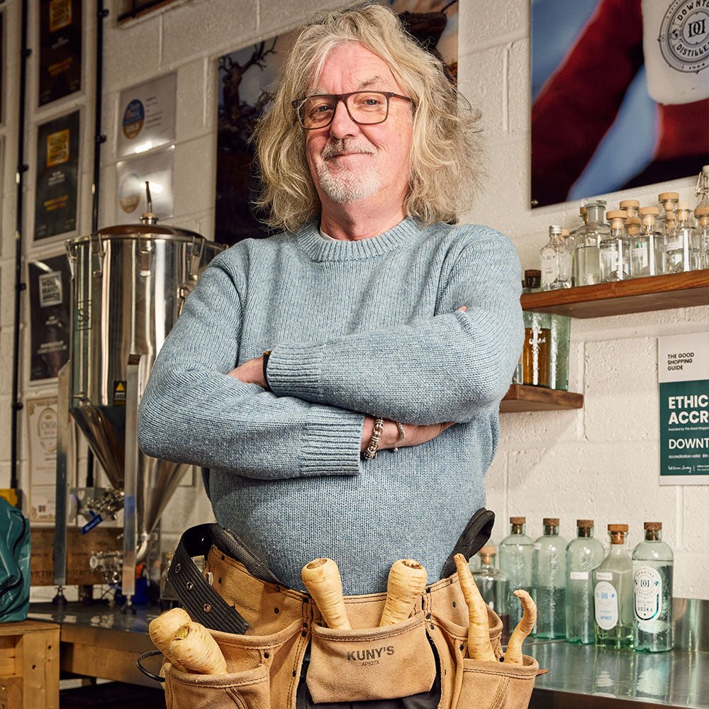 A man with gray, wavy hair and glasses stands confidently in a workshop, wearing a blue sweater and tool belt. Shelves behind him display James Gin US’s The Tupac 2 Bottle Set with Souvenir Gift Key Chain.