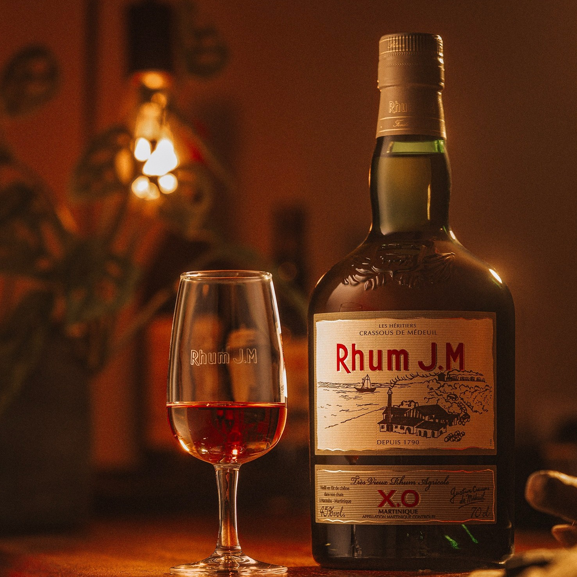 A bottle of Rhum J.M X.O., crafted by Rhum J.M and aged in re-charred Bourbon barrels, stands with a filled glass on a warmly lit table. Notes of mango and passion fruit mingle as a plant and glowing lamp soften the cozy background.
