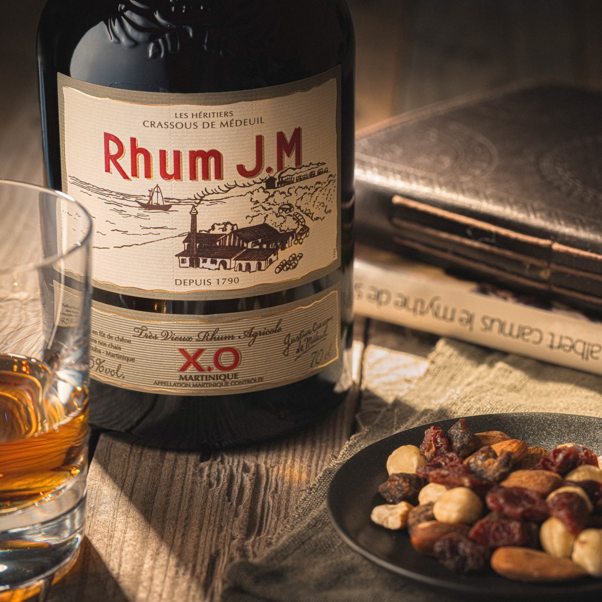 A bottle of Rhum J.M X.O. by Rhum J.M sits beside a glass of rhum, mixed nuts, dried fruit, and a closed book on a wooden table, evoking cozy elegance with notes of mango and passion fruit from re-charred Bourbon barrels.