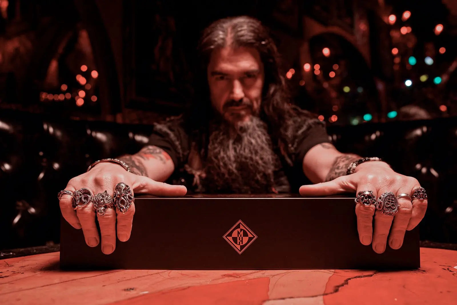 A man with long hair and a beard sits at a table, gripping a Shotgun Blast Whiskey First Edition Straight Bourbon collector’s box. He wears ornate rings as dramatic reddish lighting casts a mysterious vibe, highlighting the unique Machine Head branded product.