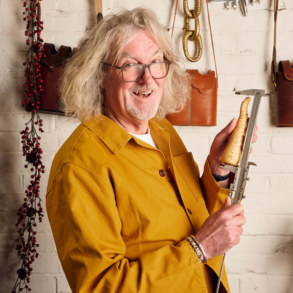 A man with gray, wavy hair and glasses smiles while measuring a parsnip, standing before a white brick wall adorned with leather bags. He wears a mustard jacket—ideal for fans of James Gin US's Six Pack Asian Parsnip & Navy Strength set.