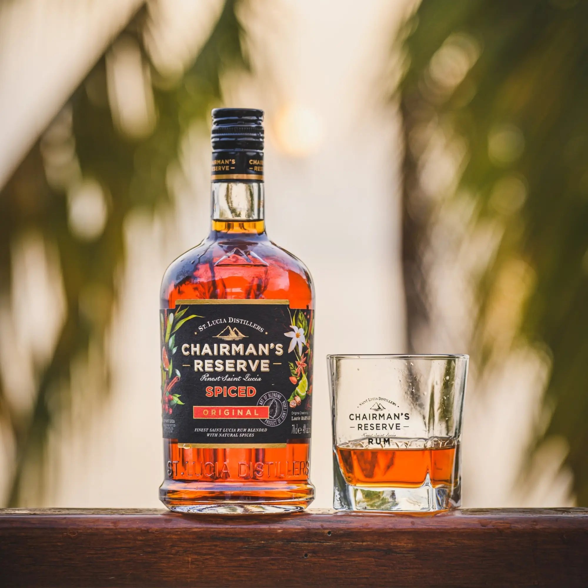 A bottle of Chairman's Reserve Spiced Original from Chairman's Reserve Rum and a glass of Caribbean rum rest on a wooden surface, framed by blurred palm leaves and sunlight in the background.