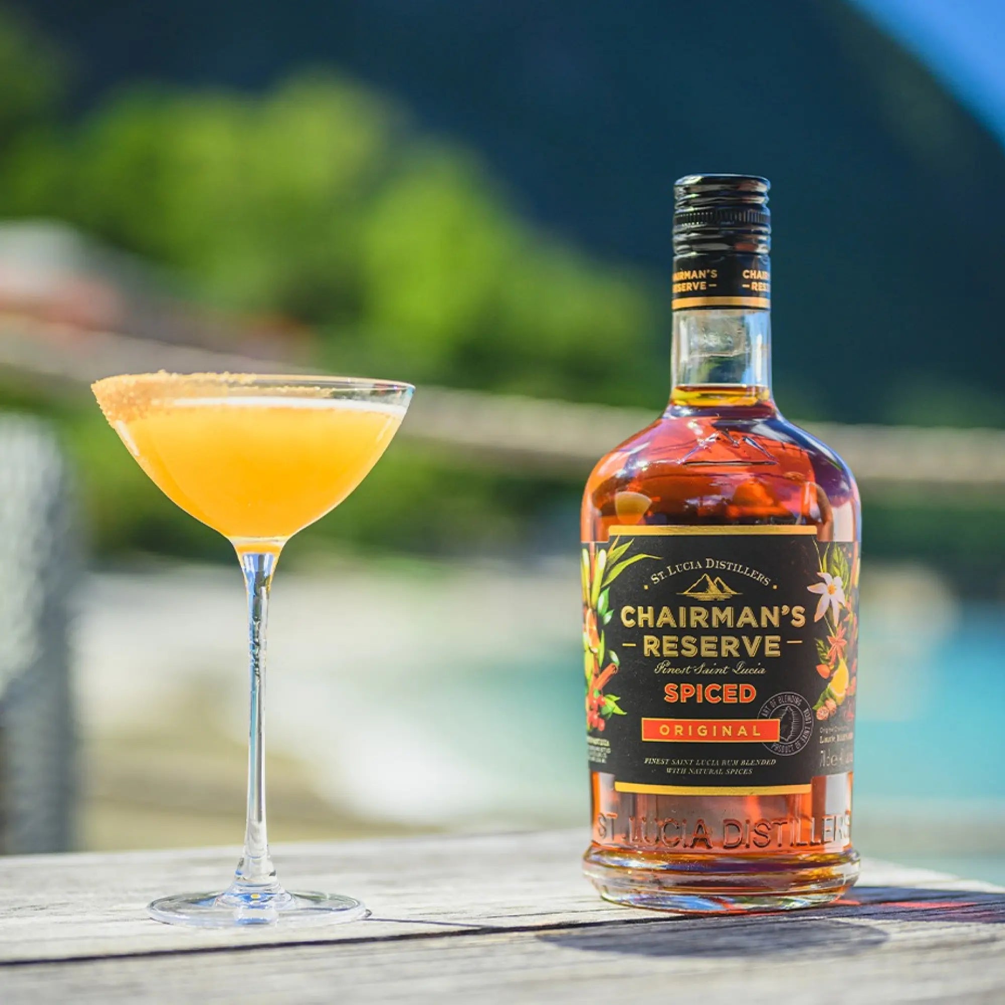 A cocktail glass with an orange drink sits beside a bottle of Chairman’s Reserve Spiced Original from Chairman’s Reserve Rum on a wooden table outdoors, with greenery and mountains blurred in the background.