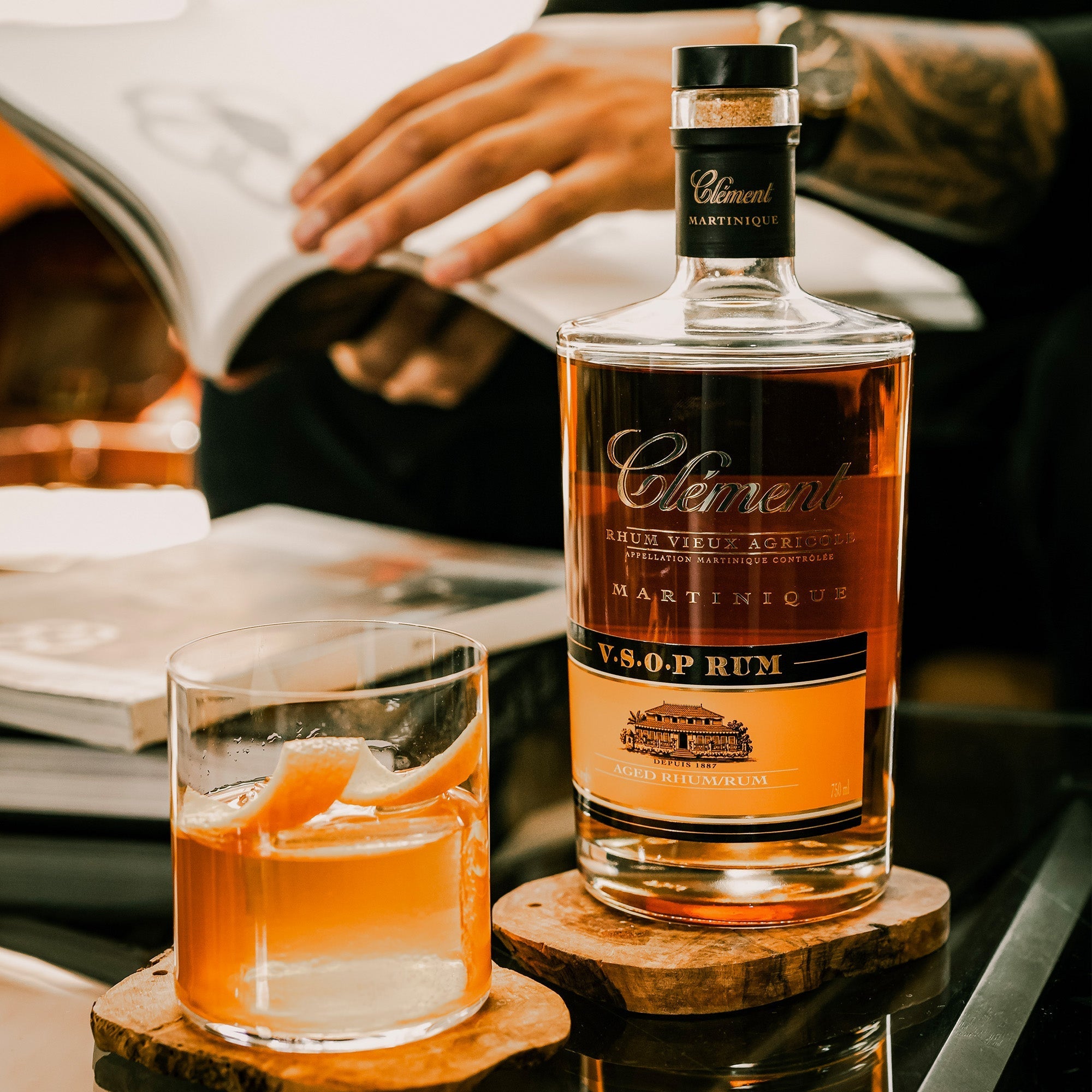 A bottle of Rhum Clément V.S.O.P. and a glass of AOC Martinique rum garnished with orange peel rest on wooden coasters, as someone reads a book in the blurred background.