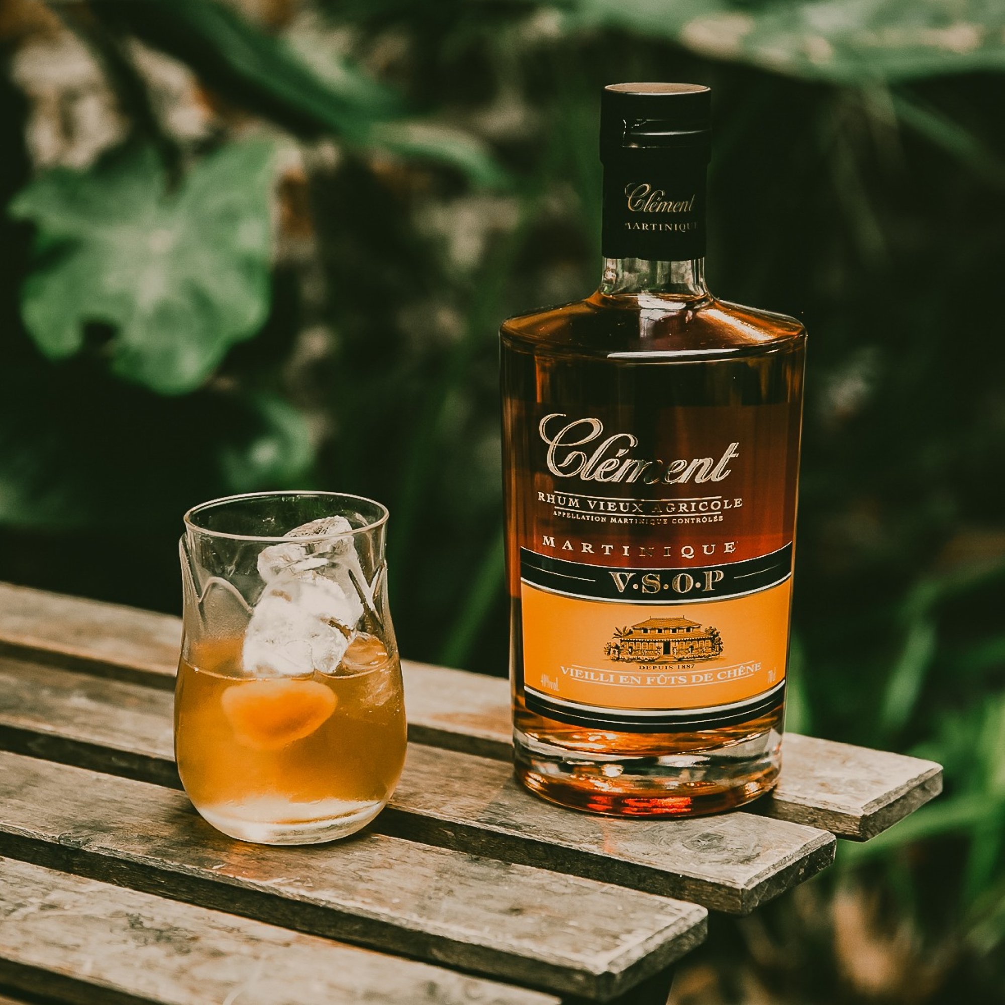 A bottle of Rhum Clément V.S.O.P. sits next to an amber cocktail with ice on a rustic wooden table outdoors, surrounded by blurred greenery in the background.