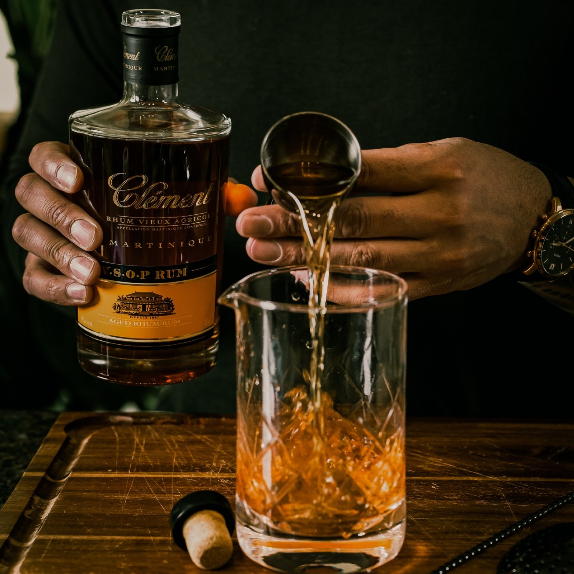 A person pours Rhum Clément V.S.O.P. from a bottle into a mixing glass on a wooden surface, with the bottle cap and a watch also visible.