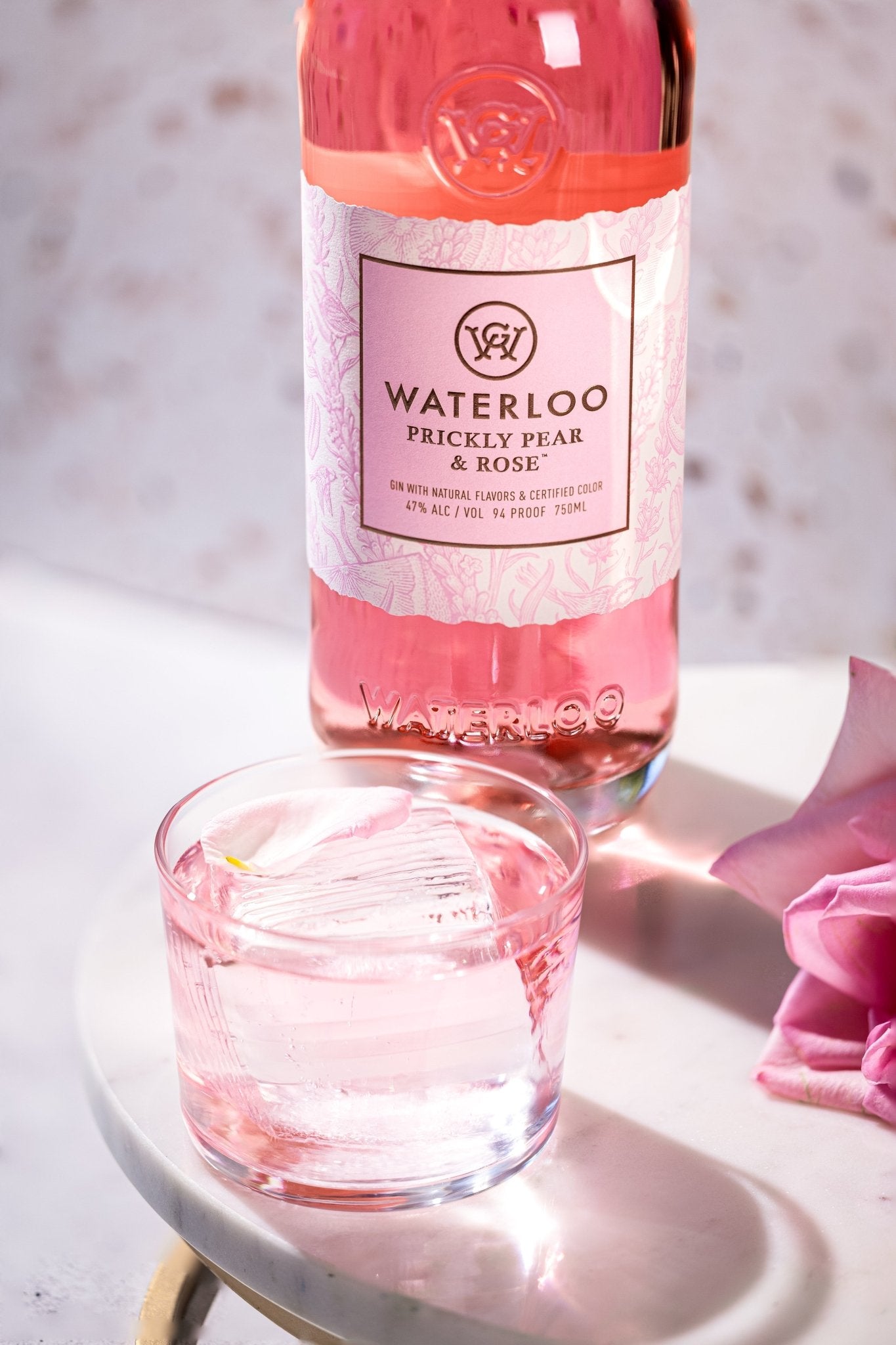 A bottle of Waterloo Gin's Prickly Pear & Rose Gin, its vibrant pink hue shining, stands behind a glass of ice and rose-tinted drink on white marble with a single pink rose petal, evoking the freshness of Texas Hill Country.